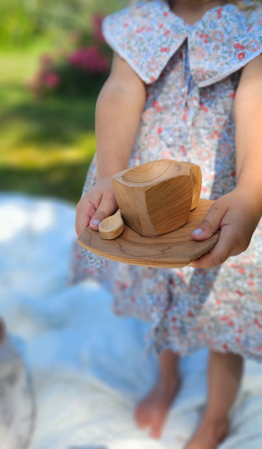 A child in a floral dress holds a small carved wooden cup and saucer outdoors on a white blanket.