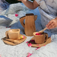 Load image into Gallery viewer, A child wearing a white patterned dress sits on a white quilted blanket outdoors, using a small wooden spoon to stir a wooden cup. Two matching wooden cups and saucers and a wooden pitcher are arranged on the blanket, with pink flower petals scattered around.
