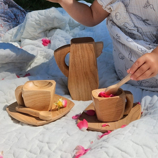 A child wearing a white patterned dress sits on a white quilted blanket outdoors, using a small wooden spoon to stir a wooden cup. Two matching wooden cups and saucers and a wooden pitcher are arranged on the blanket, with pink flower petals scattered around.